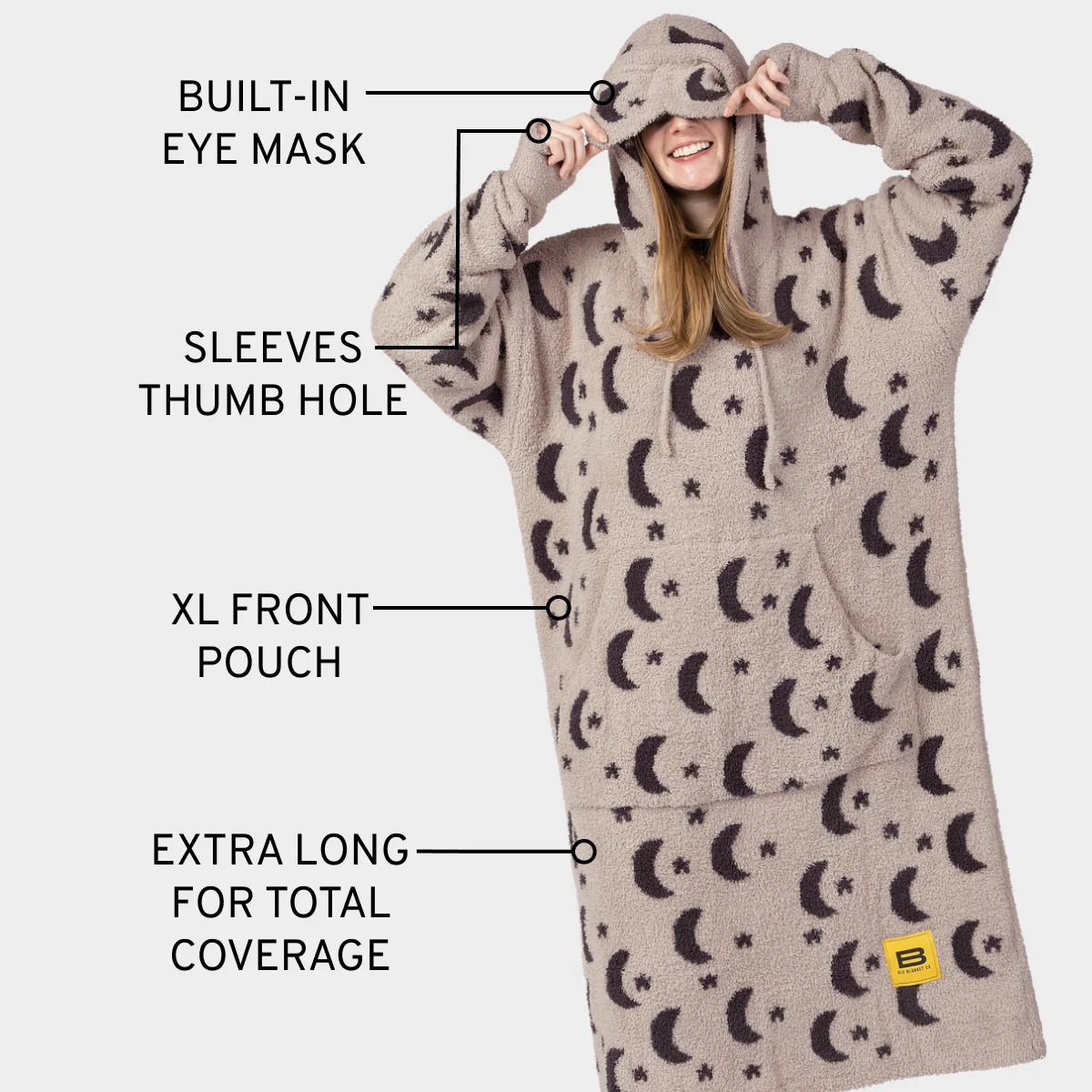 Child wrapped in cream oversized hoodie blanket with moon and star pattern, hood covering head, smiling on tan leather couch