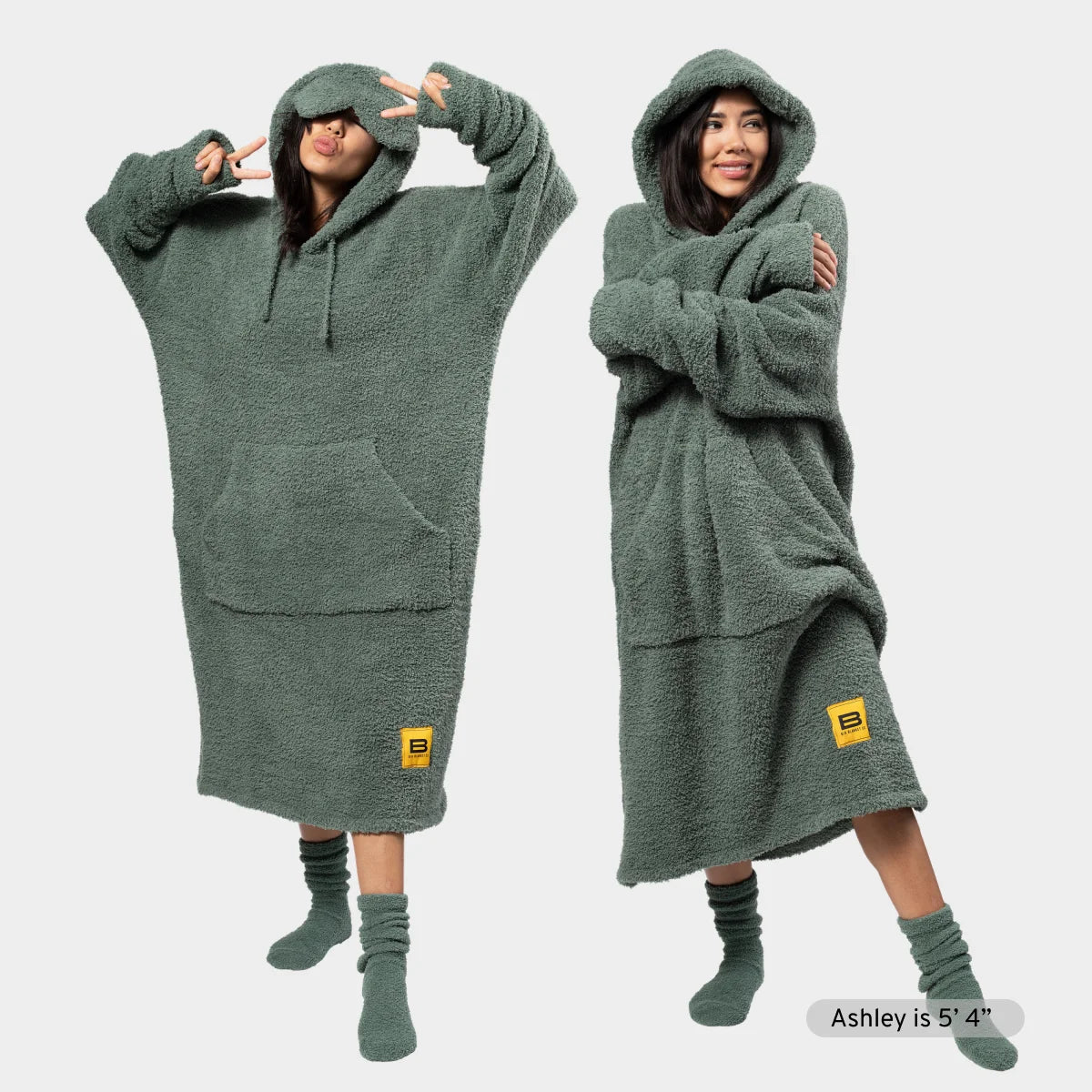 Sage green oversized wearable blanket hoodie with kangaroo pocket, shown on two models in playful poses with matching socks