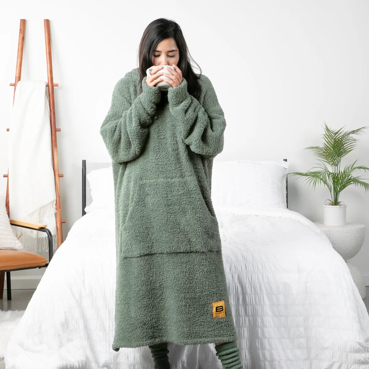 Woman wrapped in moss green oversized hoodie blanket with textured fabric, holding warm beverage in bedroom setting