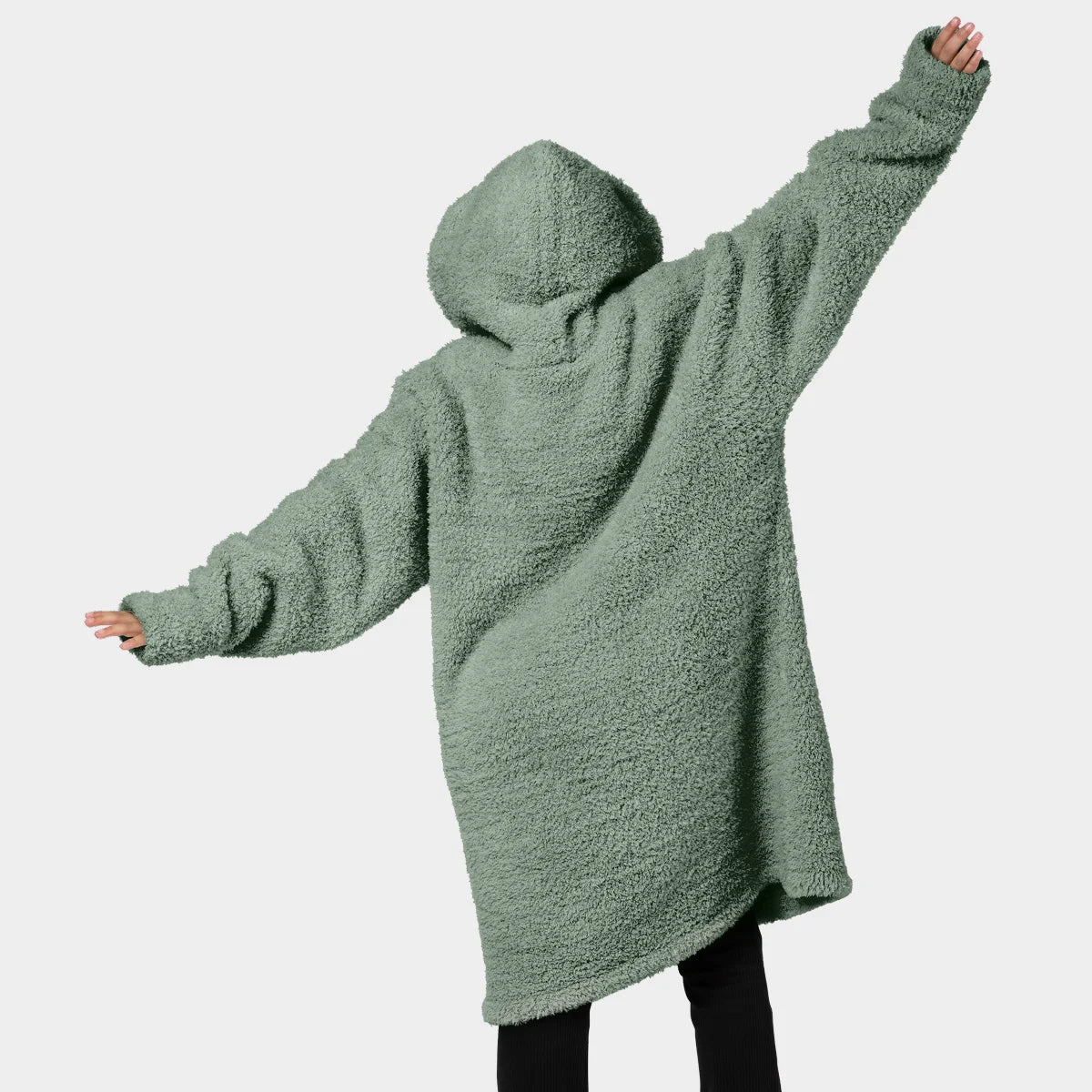 Man wearing sage green oversized hoodie blanket with kangaroo pouch, standing on bed in bedroom