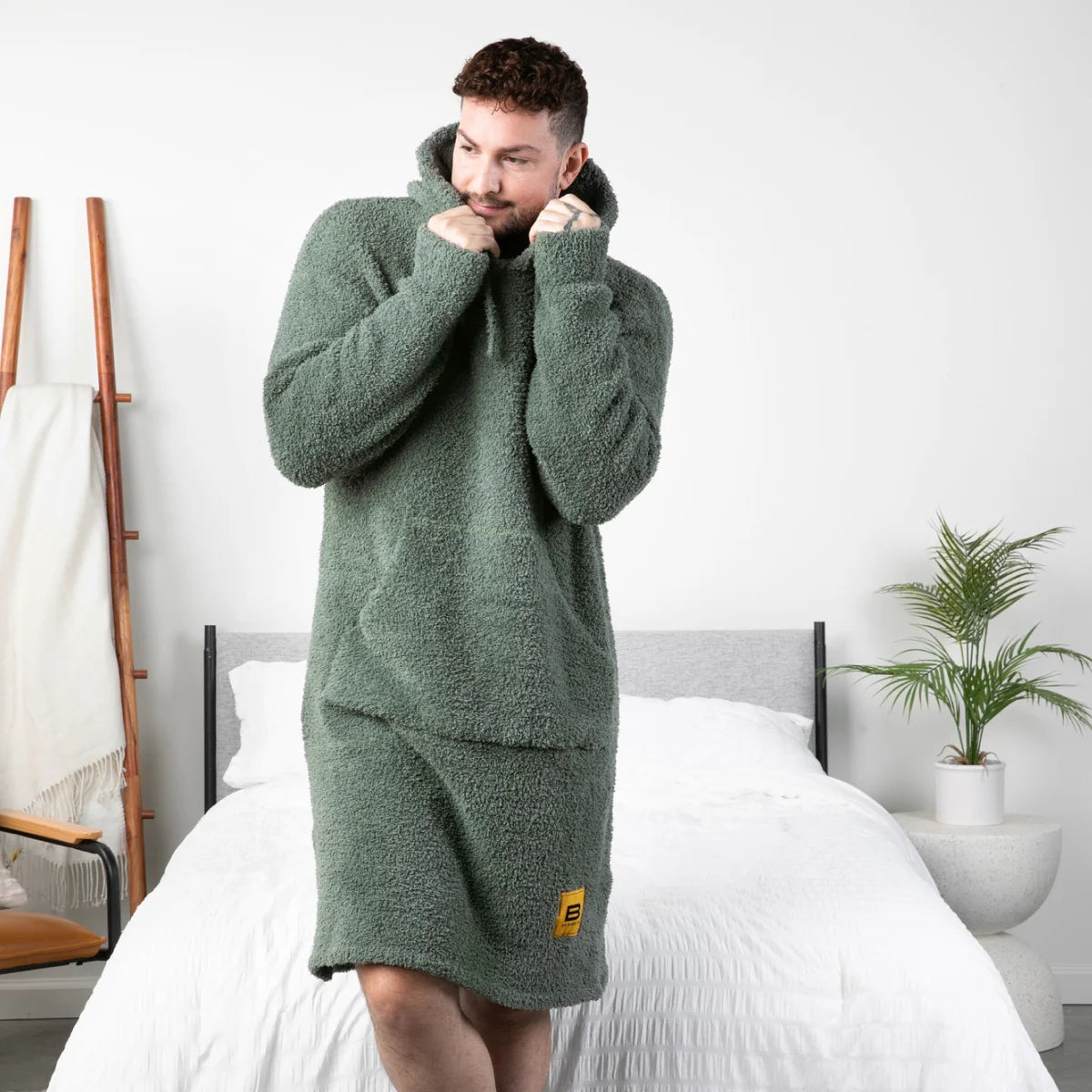 Man wearing moss green oversized hoodie blanket with hood pulled up, standing on bed in bedroom setting