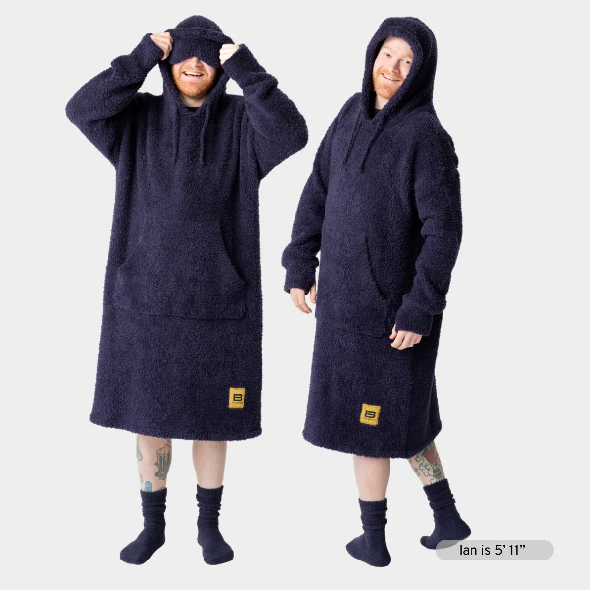 Dark navy oversized hoodie blanket worn by two models, featuring fuzzy texture, front pouch pocket, and large hood covering head