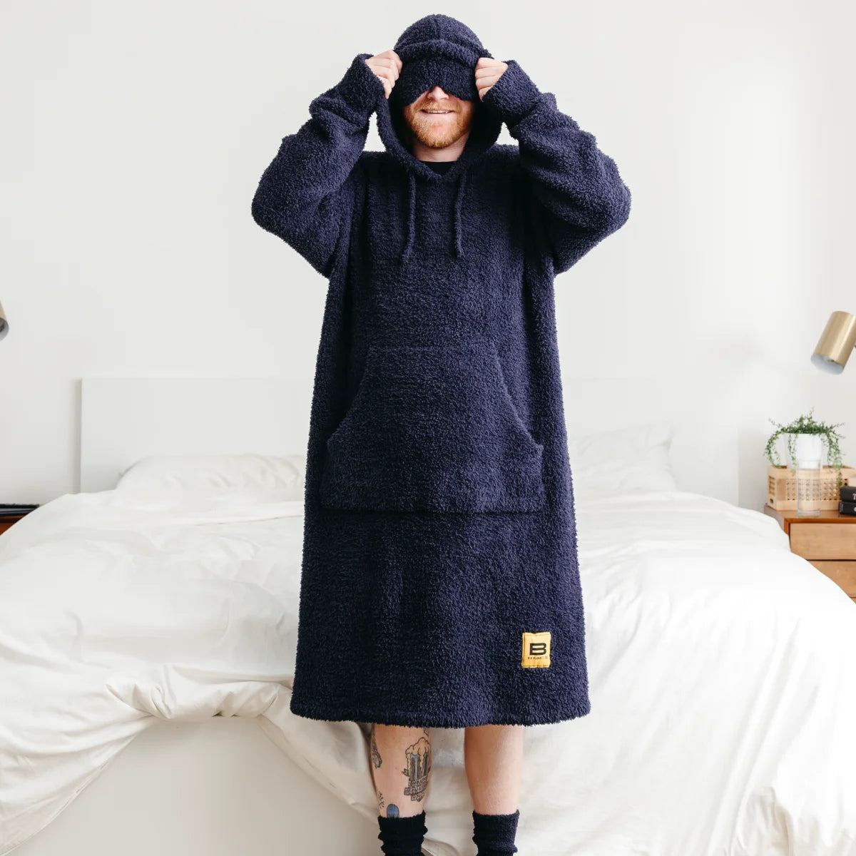 Man wearing navy oversized hoodie blanket with hood pulled over head, sitting on white bedding