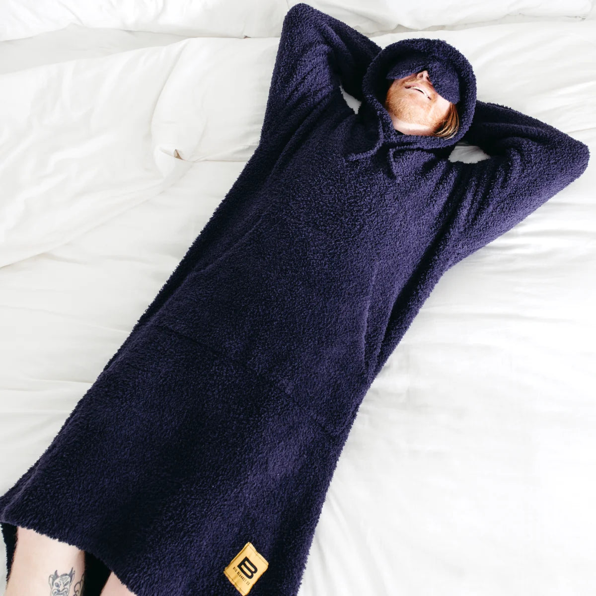 Person wrapped in dark navy oversized hoodie blanket with hood covering head, lying on white bedding