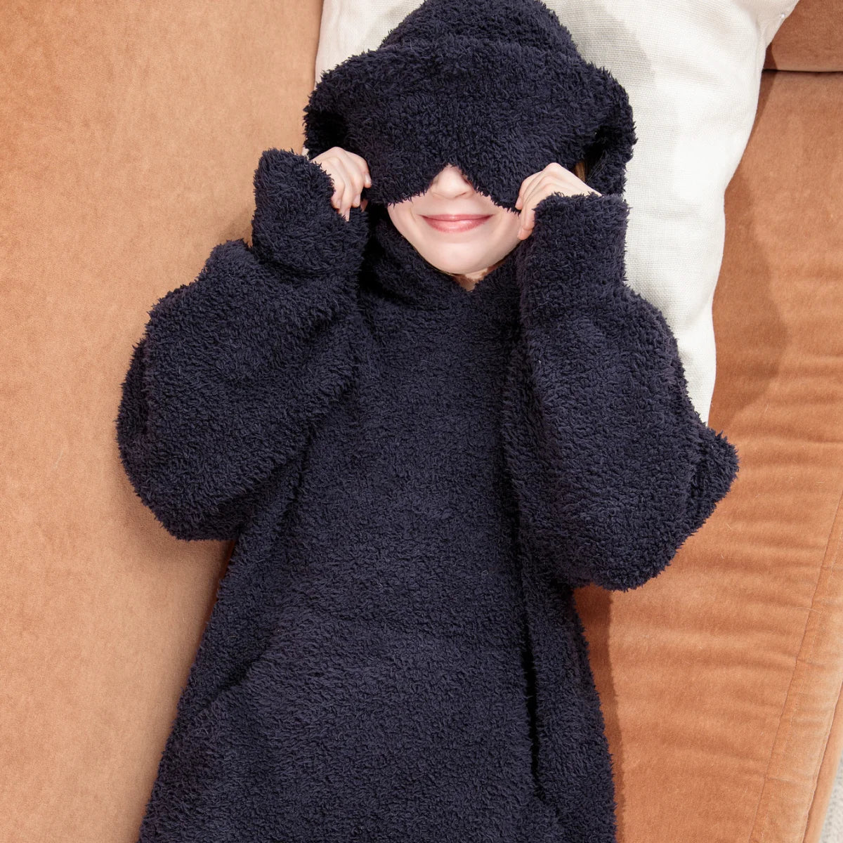 Person wrapped in navy plush hoodie blanket with hood on white bedding