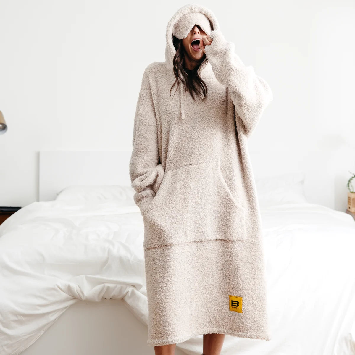 Woman wearing oversized oatmeal hoodie blanket with deep pockets and hood, sitting on bed with white bedding