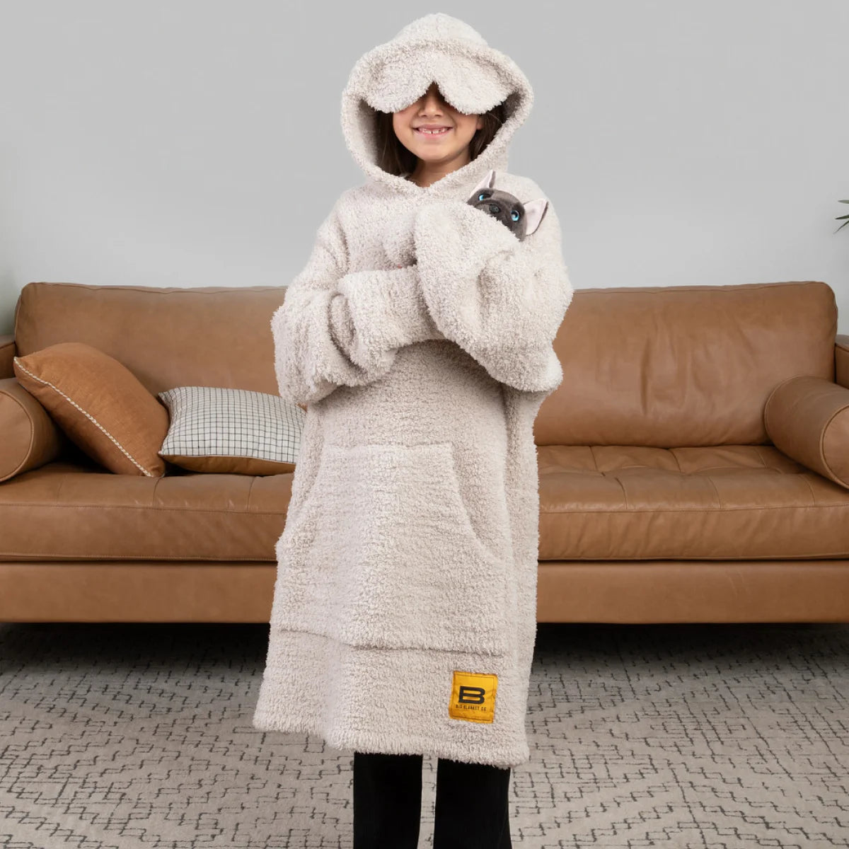 Woman wearing cream oversized hoodie blanket with hood up, standing on bed with white bedding