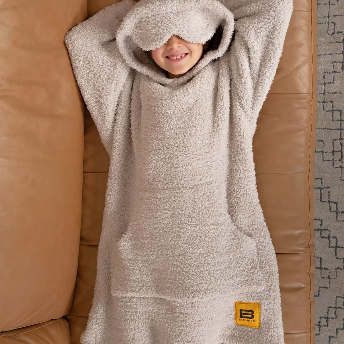 Child wrapped in oatmeal Premier Plush hoodie blanket on white bedding, smiling with hood pulled over head