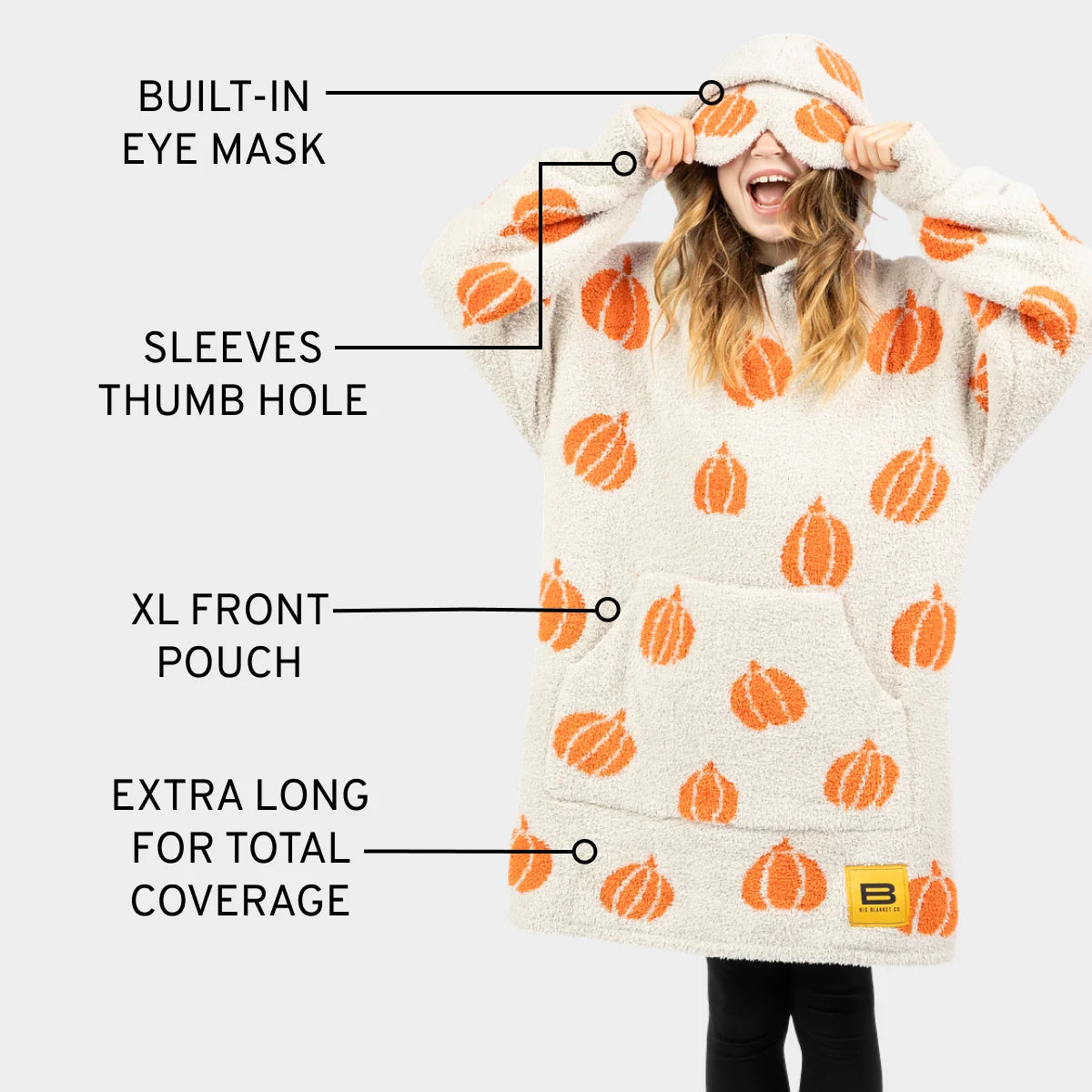 Woman wearing cream oversized hoodie blanket with orange pumpkin pattern, built-in eye mask and thumb holes, demonstrating features