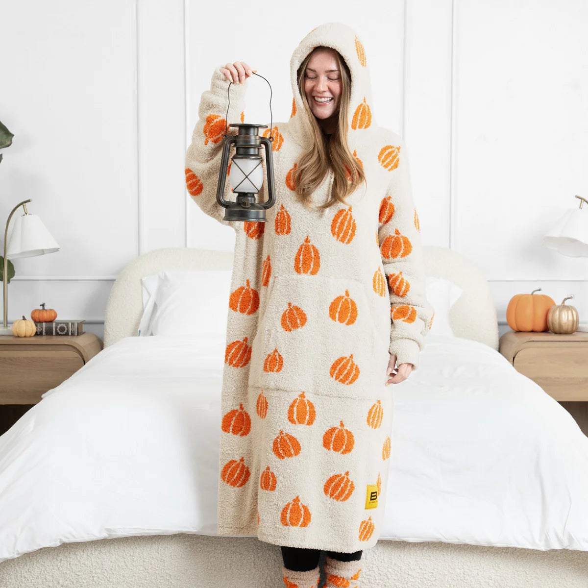 Woman wrapped in cream oversized hoodie blanket with orange pumpkin pattern, holding lantern on bed