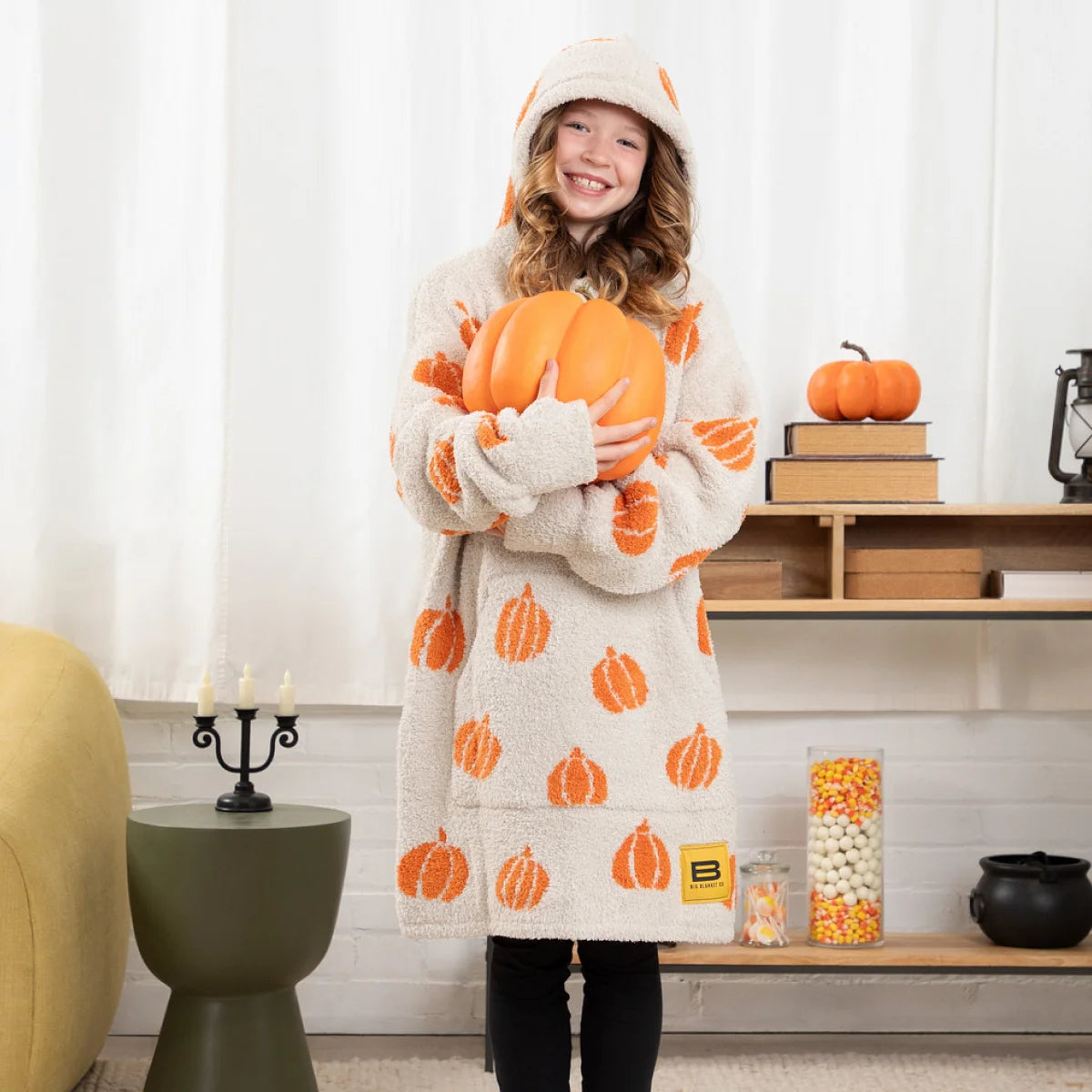 Woman wrapped in cream oversized blanket with orange pumpkin pattern and hood, holding lantern on bed
