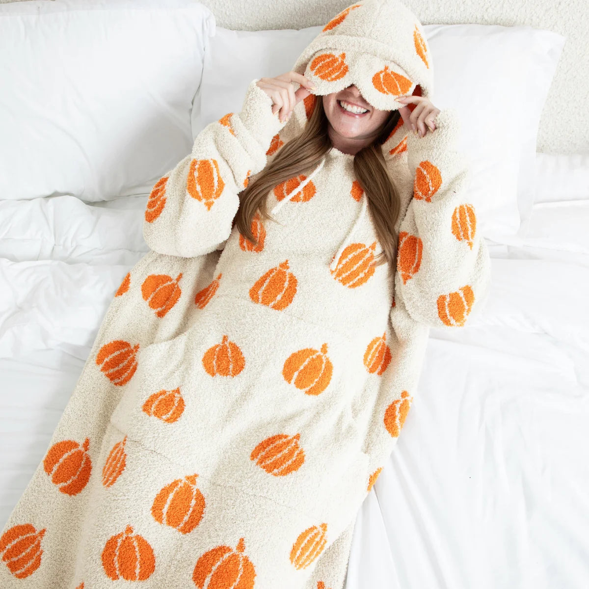 Cream oversized hoodie blanket with orange pumpkin pattern, worn by smiling woman on bed with hood covering eyes