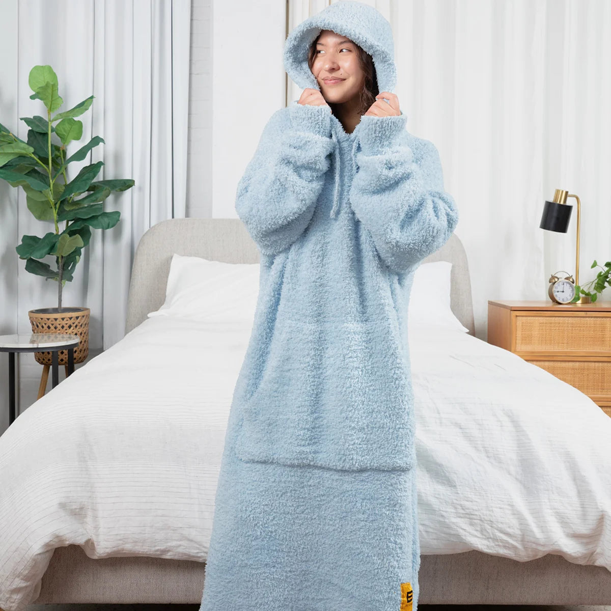 Woman wearing light blue oversized hoodie blanket with hood on bed, showing plush texture and full-length coverage