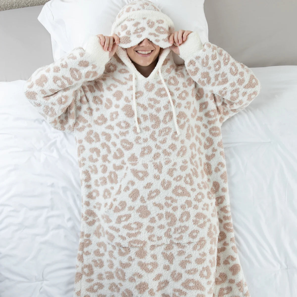 Person wrapped in cream and taupe leopard print oversized hoodie blanket with matching sleep mask on bed