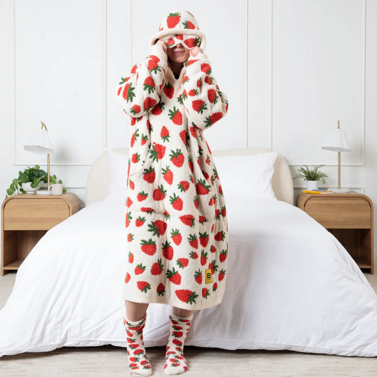 Person wearing cream Premier Plush™ Hoodie in strawberry print with matching hood and socks, standing on bed