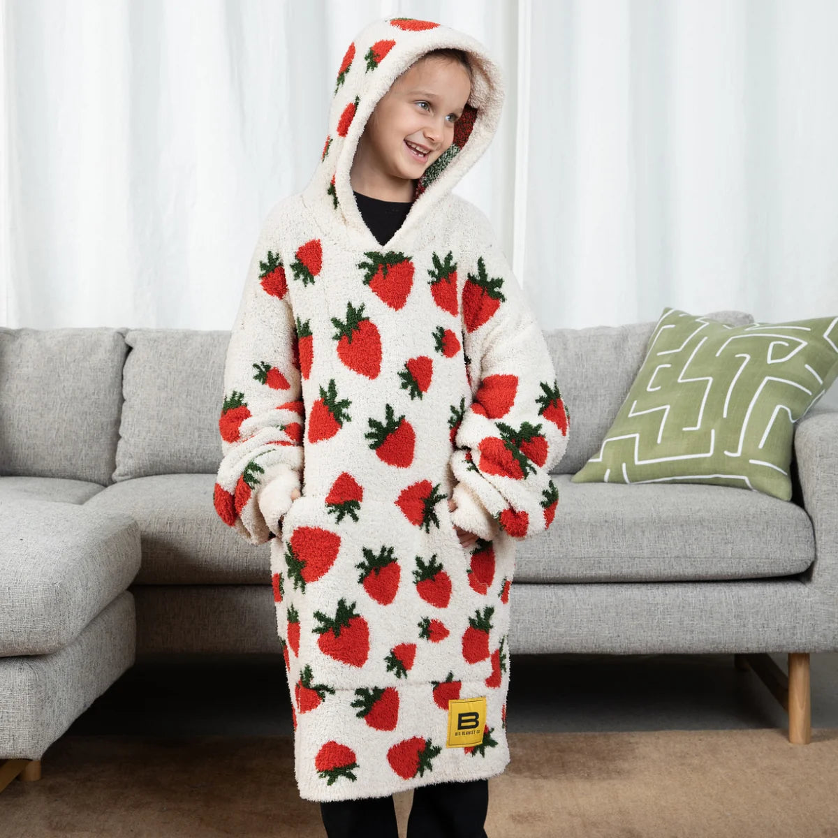 Person wearing cream hoodie blanket with red strawberry pattern and matching hood, standing on bed