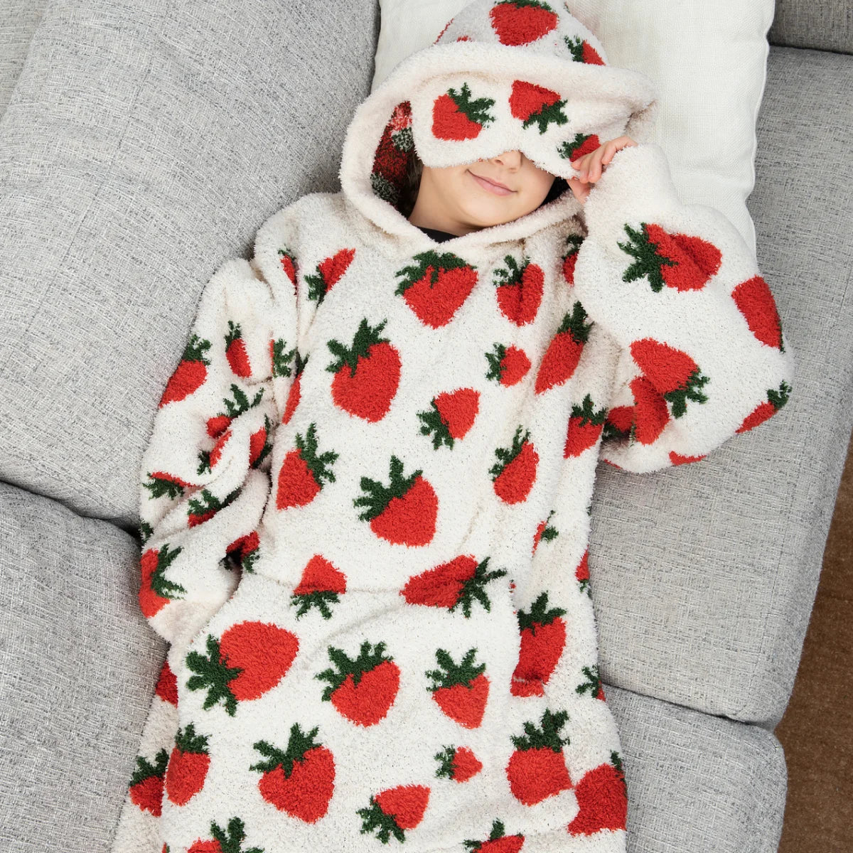 Smiling child wrapped in cream Premier Plush hoodie blanket with red strawberry and green leaf pattern