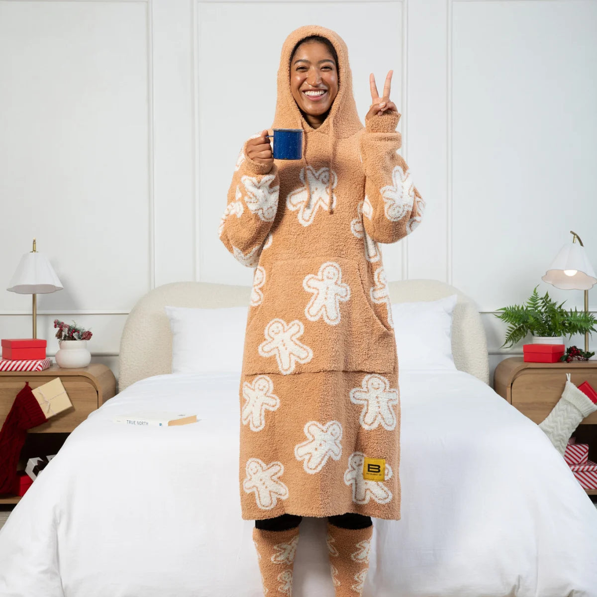 Woman wearing tan gingerbread-patterned hoodie blanket with white icing details, holding blue mug on bed