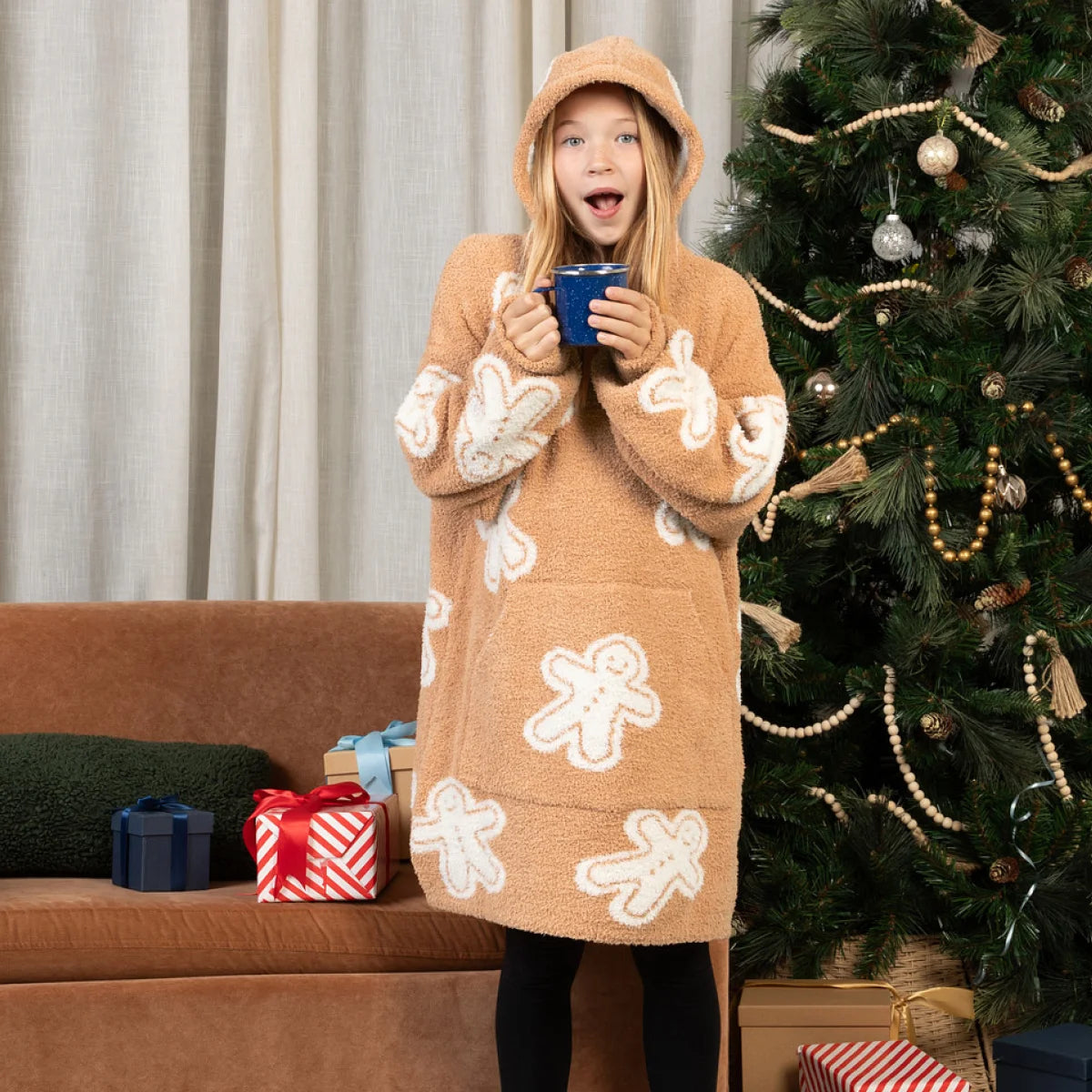 Woman wearing tan oversized hoodie blanket with white gingerbread pattern, holding blue mug on bed