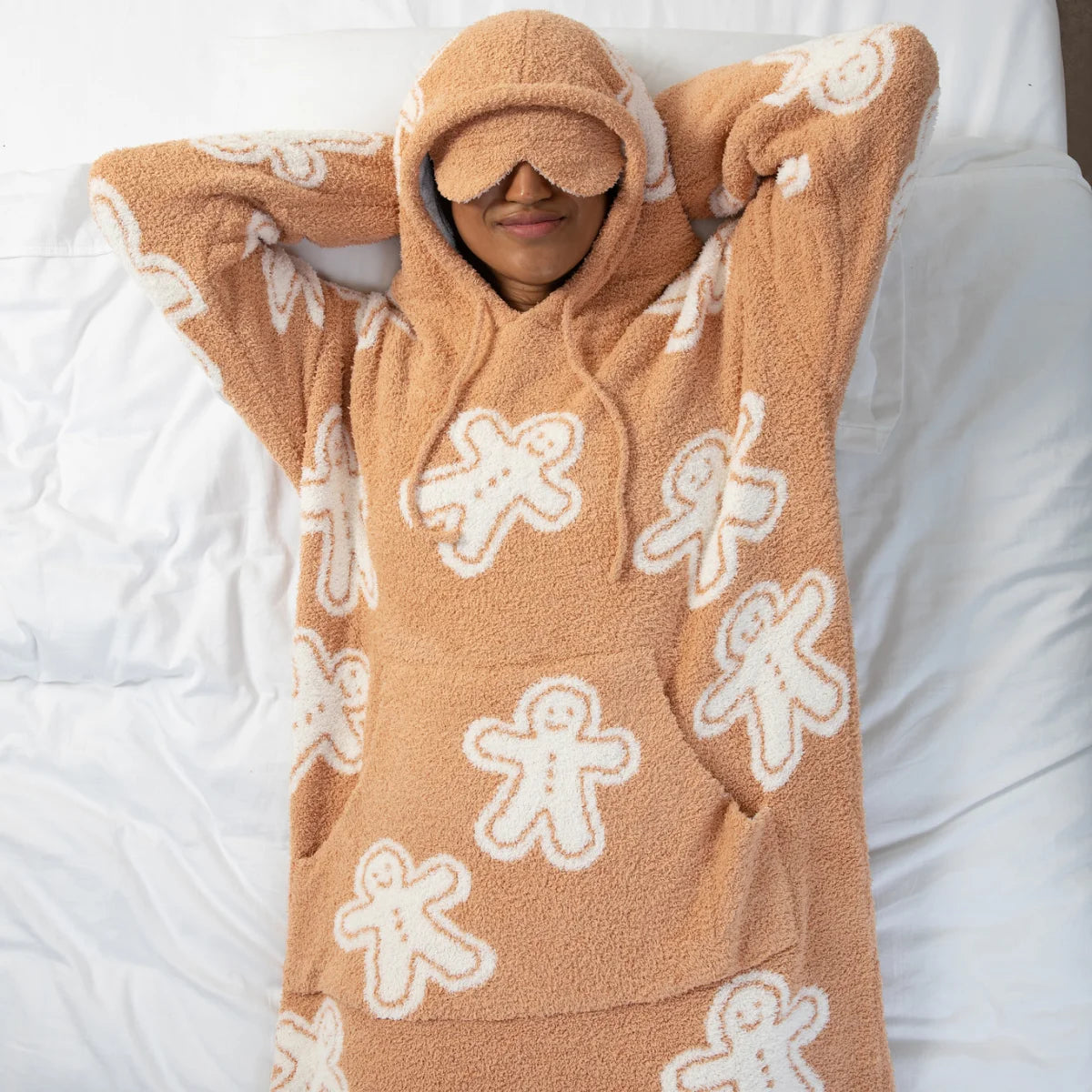 Tan Premier Plush Hoodie with gingerbread man pattern, modeled by person wearing matching eye mask on white bedding