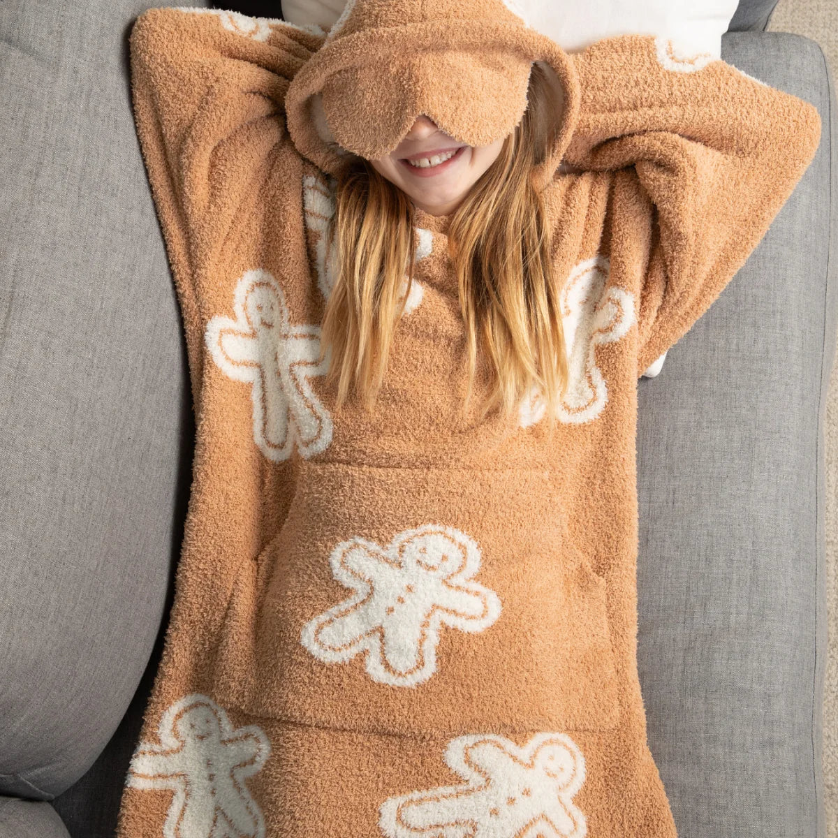 Tan oversized hoodie blanket with gingerbread man pattern, person wrapped wearing matching eye mask on white bedding