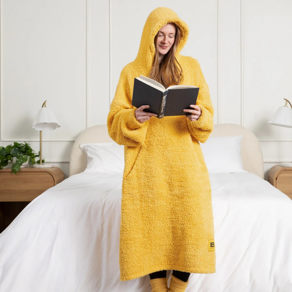 Woman wearing sunflower yellow oversized blanket hoodie with attached hood, reading on bed with white sheets