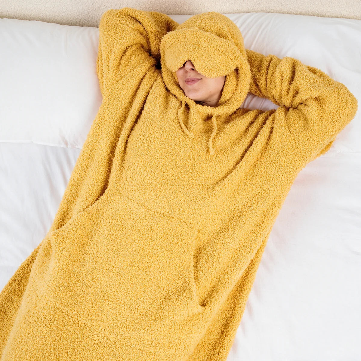 Child wrapped in yellow oversized hoodie blanket with hood up, holding turquoise bowl, sitting on gray sofa with green pillows