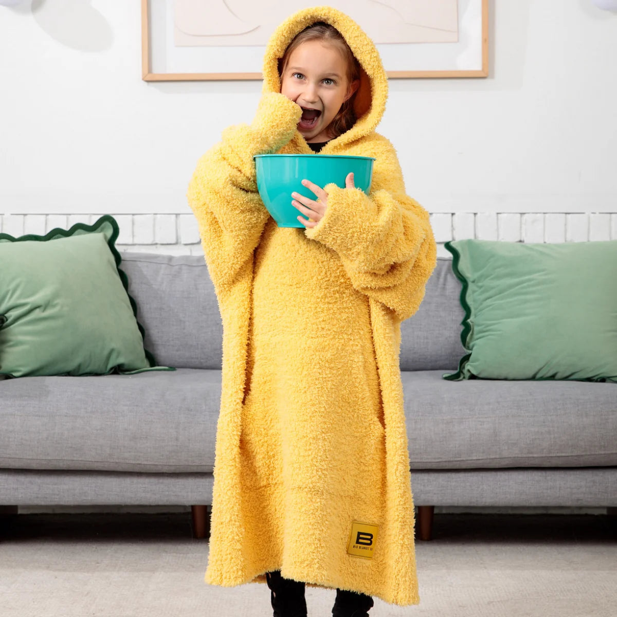 Child wrapped in yellow oversized hoodie blanket with hood up, holding turquoise bowl, sitting on gray couch with green pillows