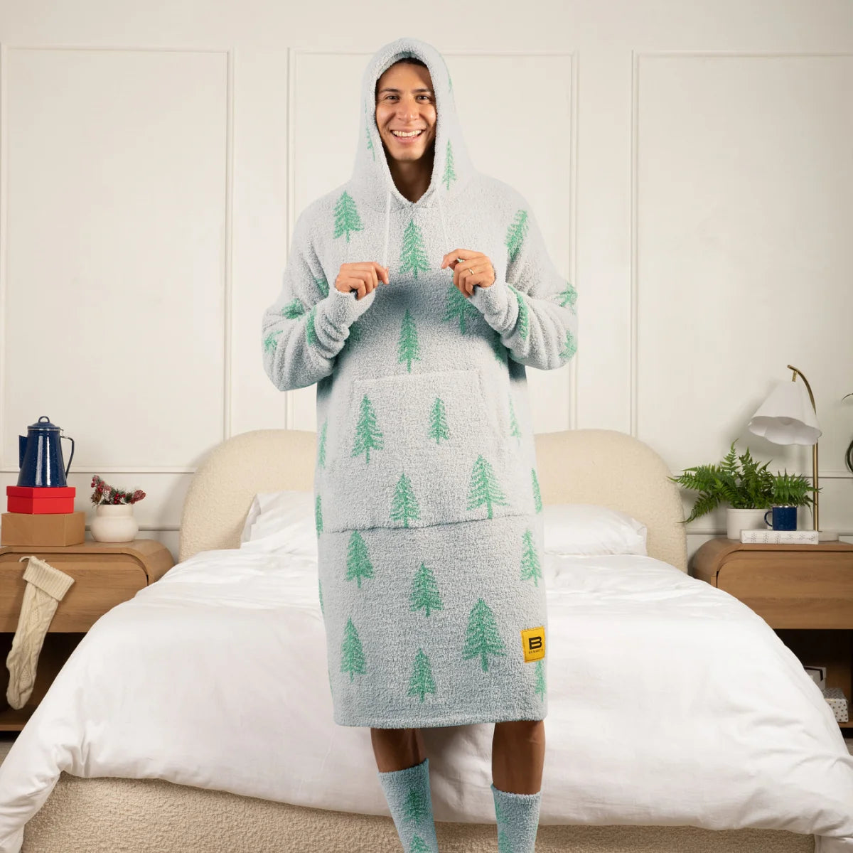 Person wearing gray oversized hoodie blanket with green pine tree pattern and matching socks, standing on bed