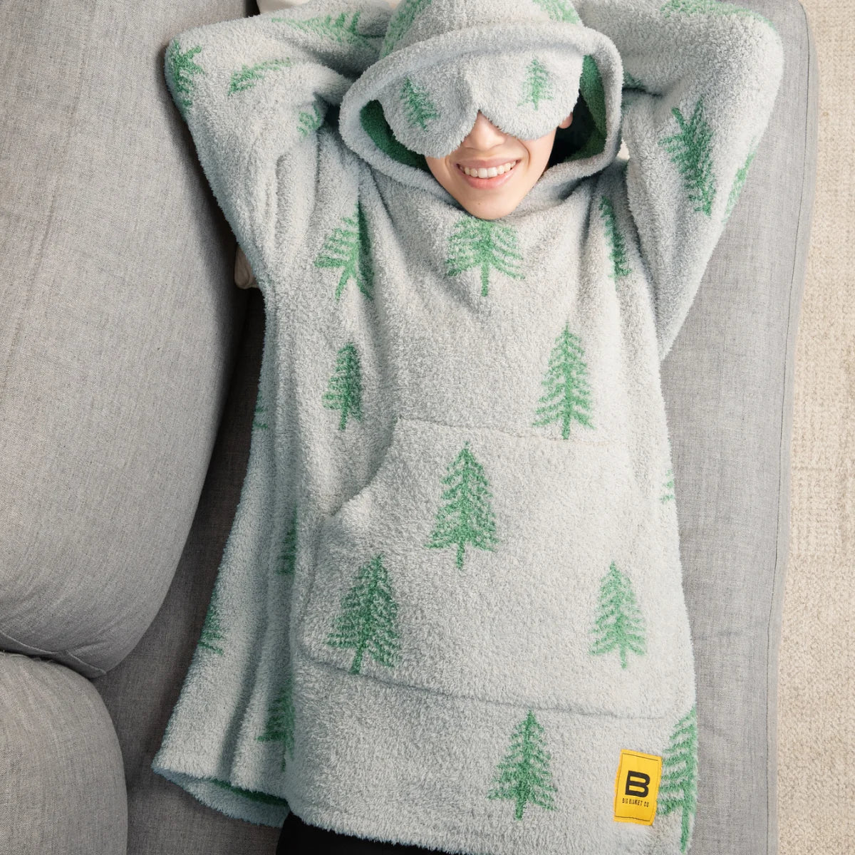 Person wrapped in oversized cream blanket with green tree pattern, hood pulled over head, smiling on white bedding
