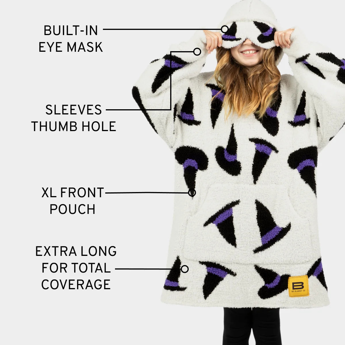 Woman wearing cream oversized blanket hoodie with purple and black witch hat pattern, built-in eye mask, sleeves with thumb holes, and XL front pouch