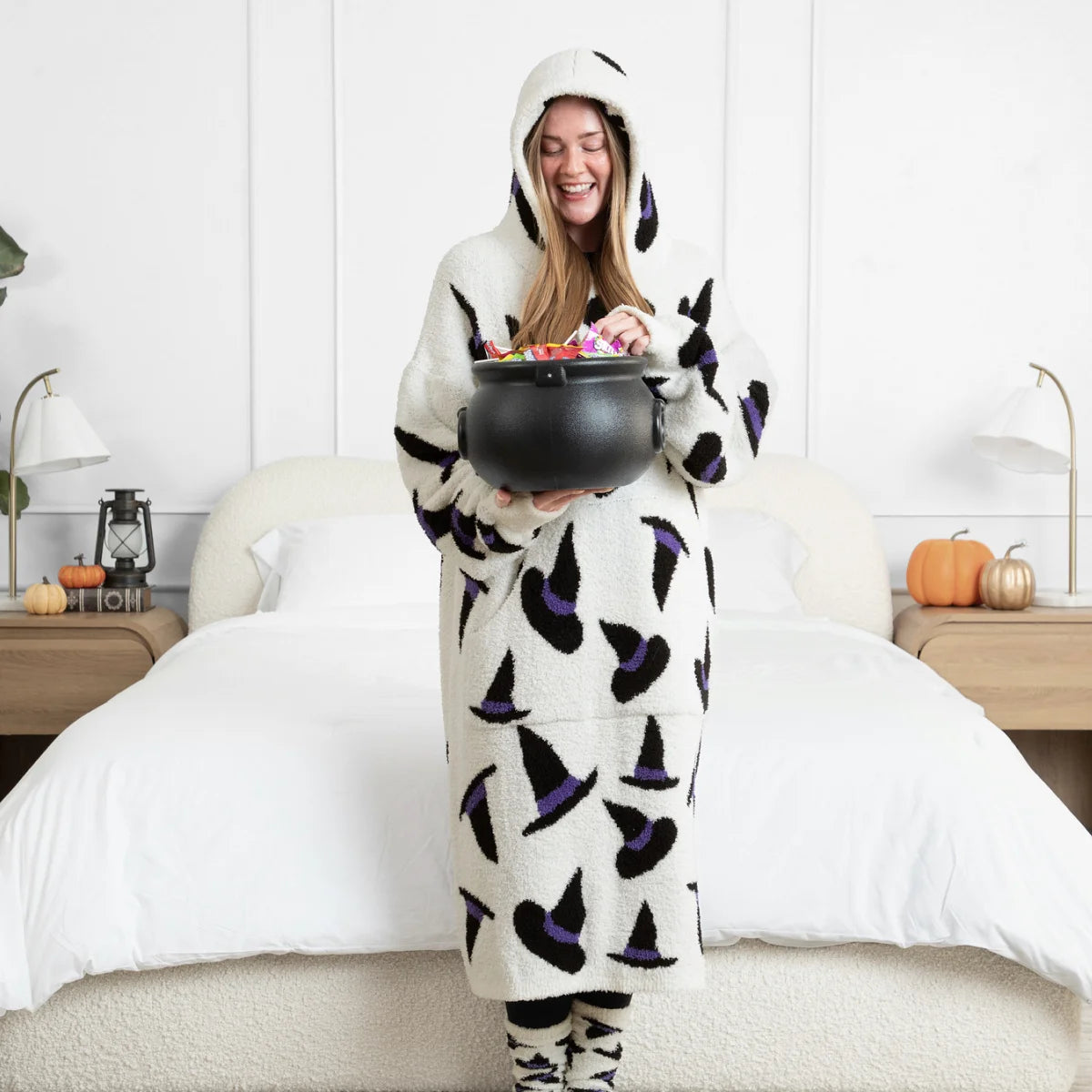 Woman wrapped in oversized cream hoodie blanket with black and purple witch hat pattern, holding cauldron, standing on bed