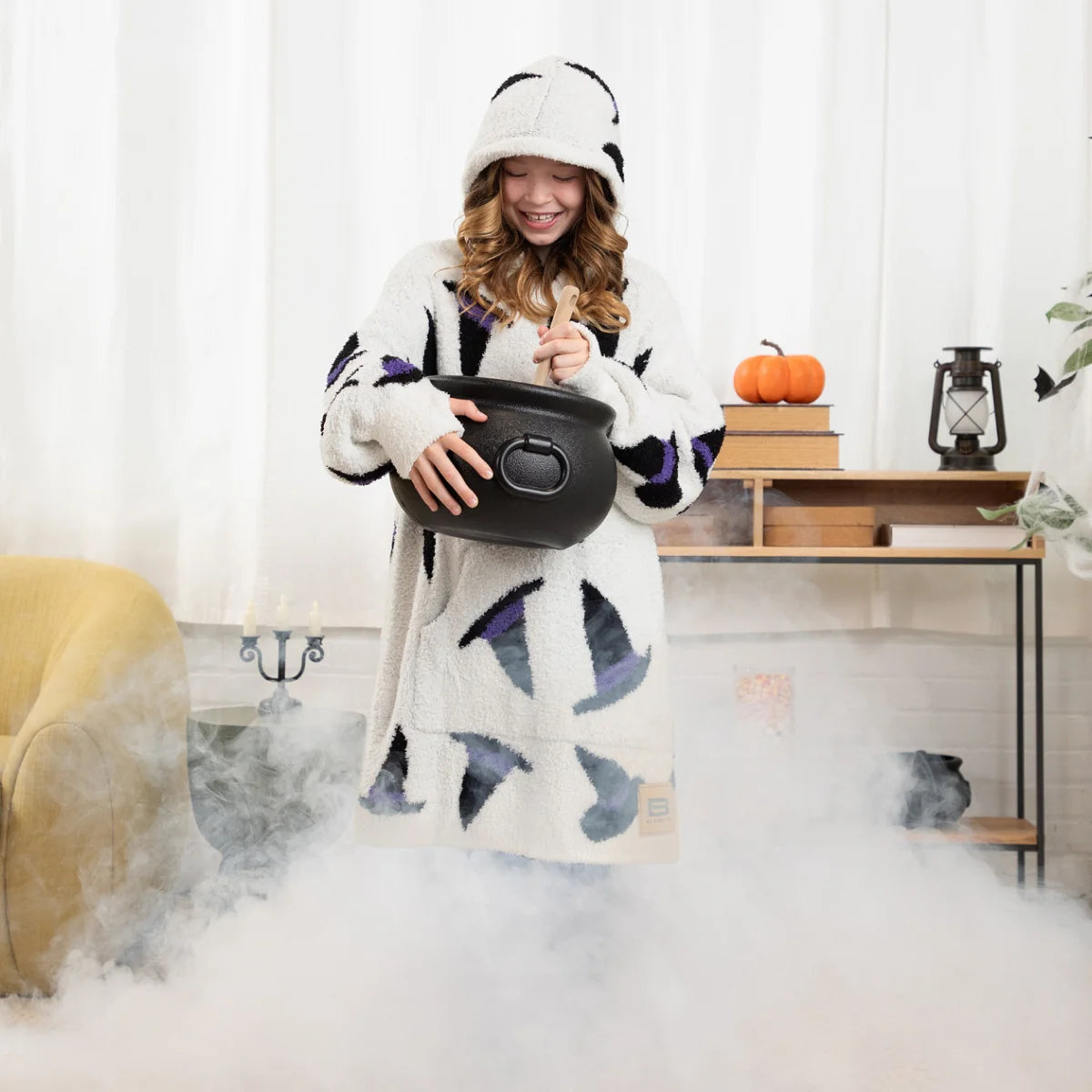 Woman wrapped in cream oversized blanket with black and purple witch hat pattern, wearing matching hoodie, holding cauldron on bed