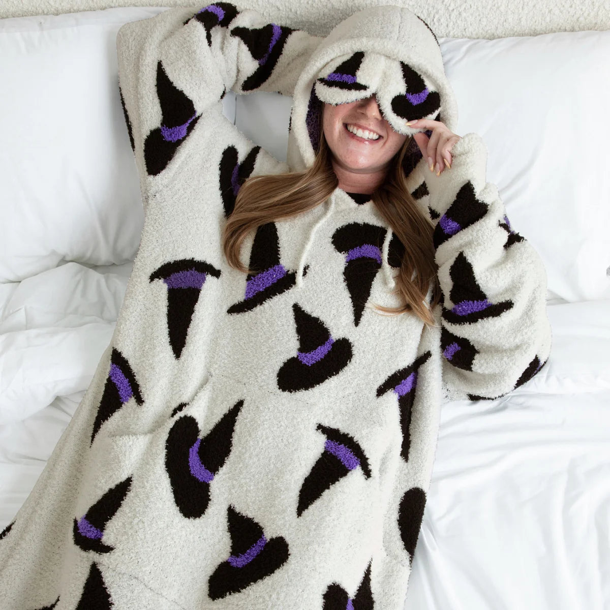 Cream oversized hoodie blanket with black and purple eye pattern, woman wearing on bed with hood up covering eyes
