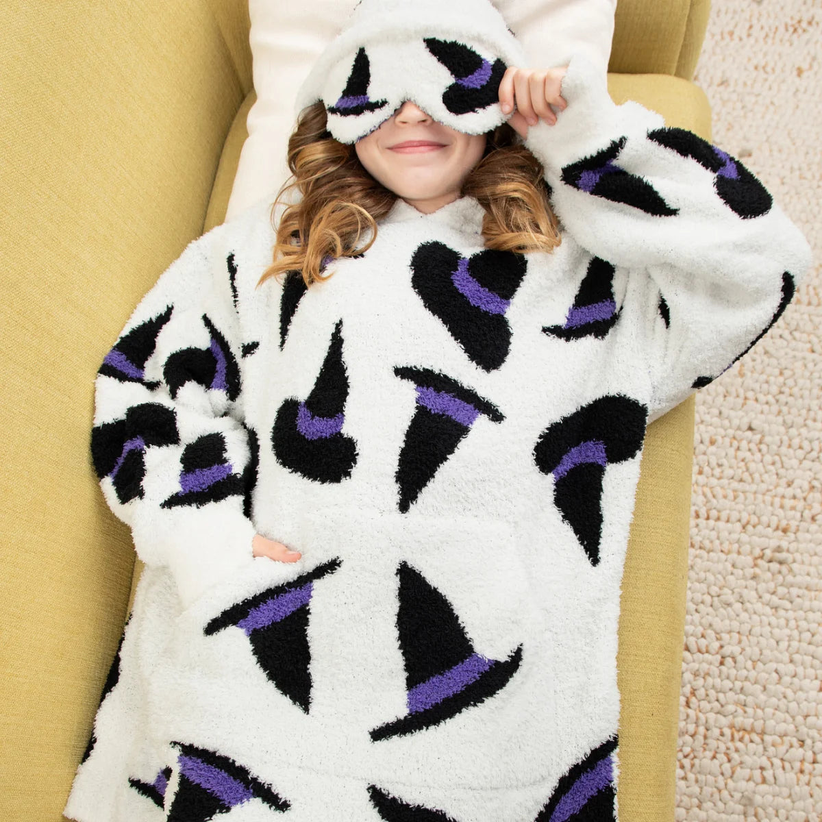 Cream oversized hoodie blanket with black and purple witch eye pattern, worn by smiling woman on bed