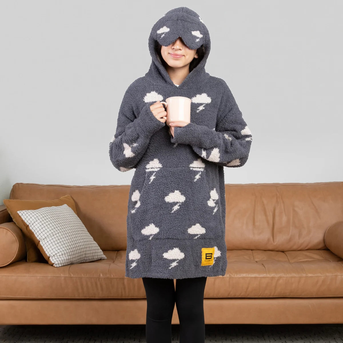 Child wearing charcoal hoodie blanket with cloud and lightning pattern, matching eye mask, holding mug on tan couch