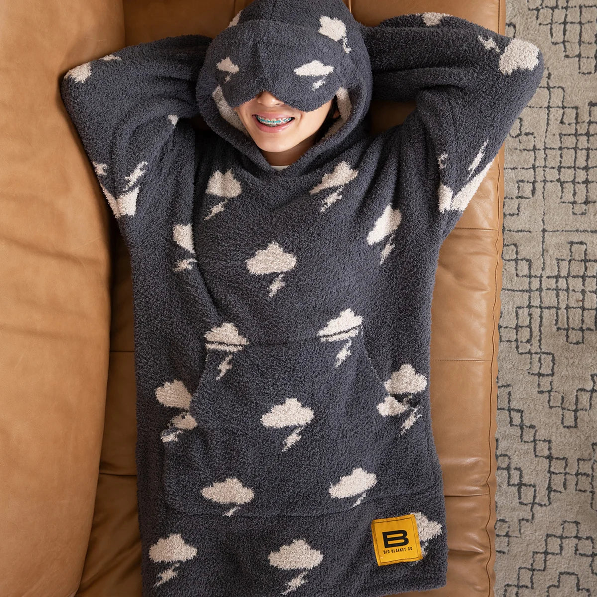 Charcoal oversized kids hoodie with white cloud and lightning bolt pattern, featuring attached hood with eye mask, worn by smiling child