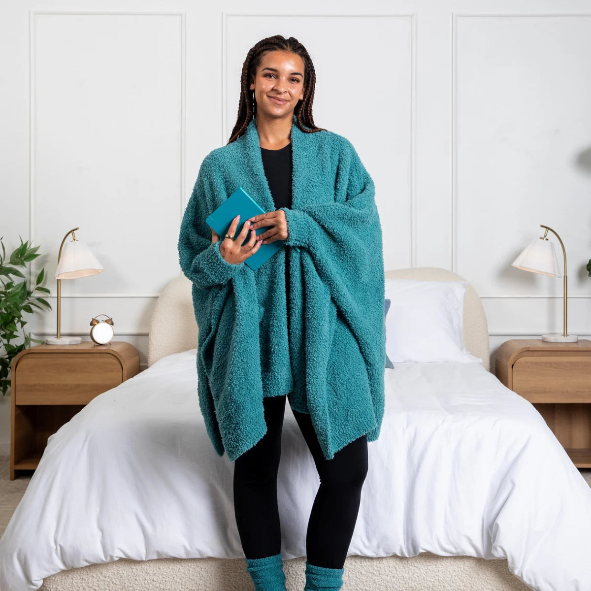 Woman wearing teal oversized plush wrap draped over shoulders on bed with white bedding