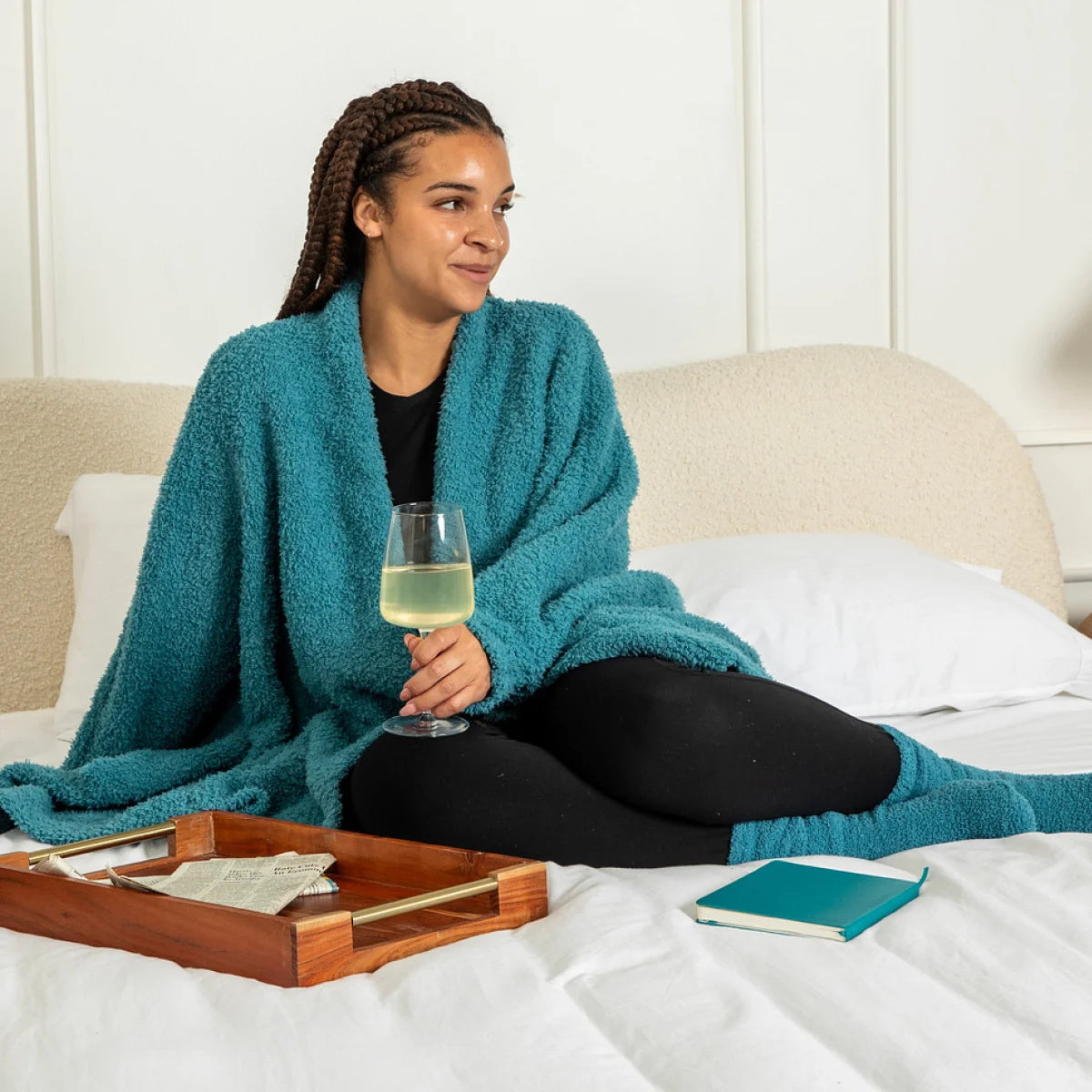 Woman wrapped in teal oversized blanket sitting on bed with wine glass and book