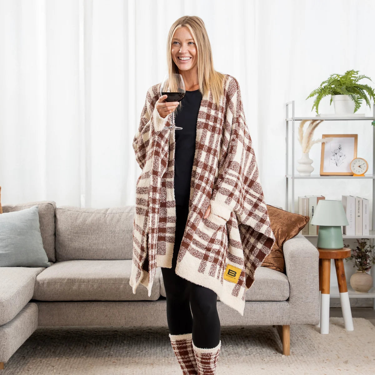 Woman wearing burgundy and cream plaid oversized blanket wrap while holding wine glass in modern living room