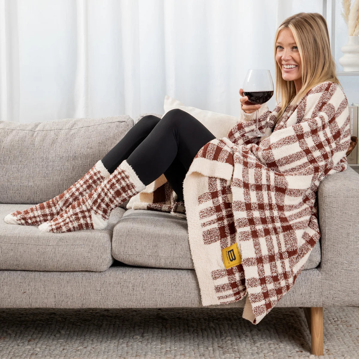 Woman wrapped in rust and cream plaid oversized blanket on couch, holding wine glass, wearing matching socks