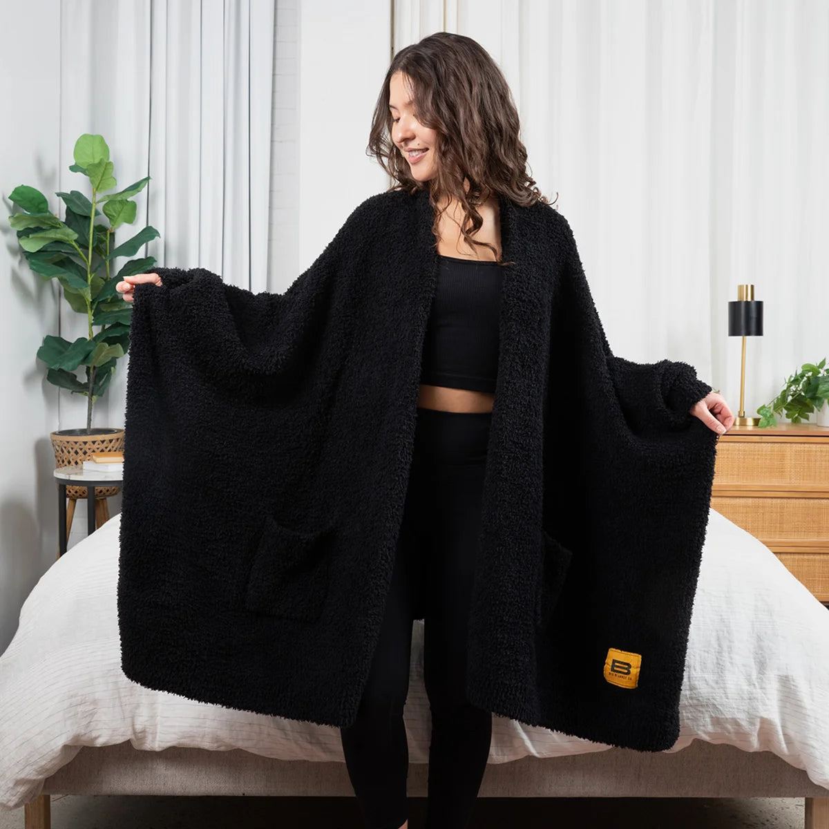 Woman wearing black oversized blanket wrap draped over shoulders on bed in modern bedroom