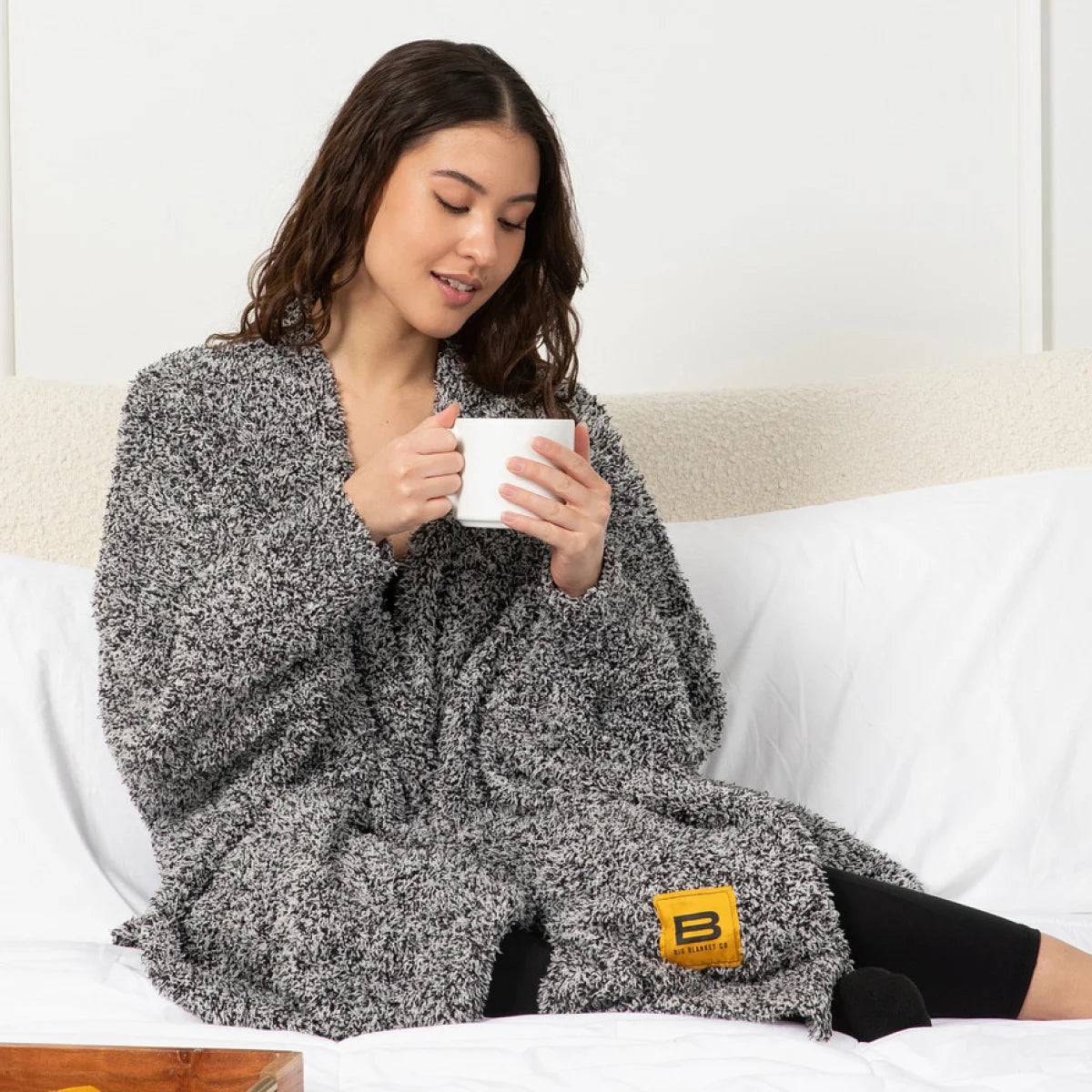 Woman wrapped in charcoal gray oversized blanket sitting on bed holding mug, Premier Plush Wrap Black Heather