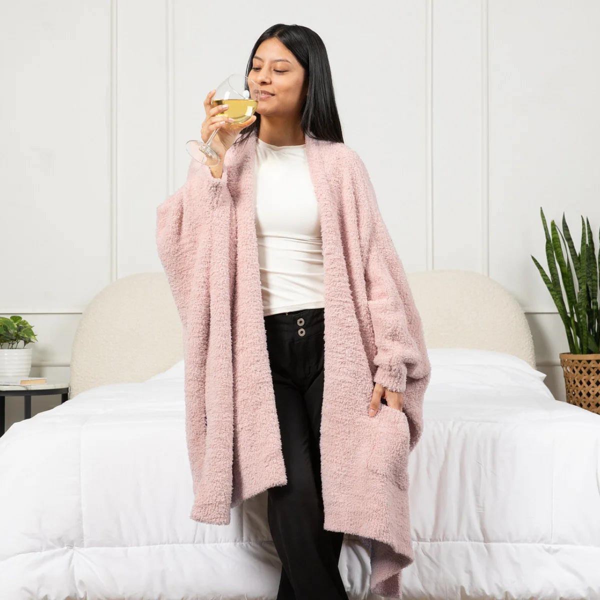 Woman wearing blush pink oversized blanket wrap on bed, drinking wine