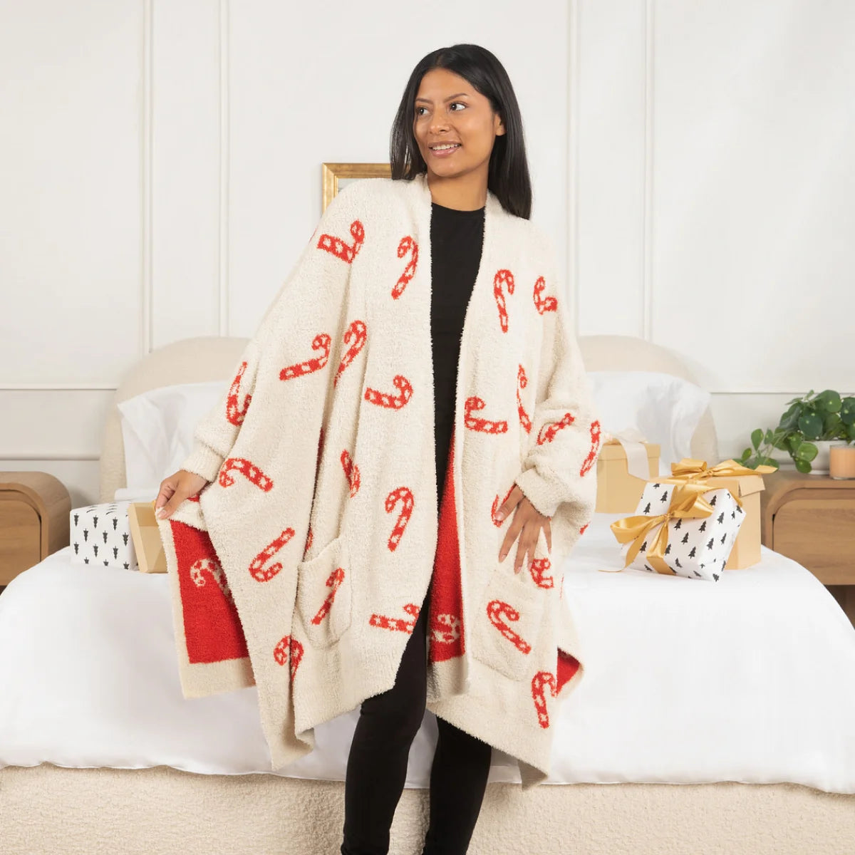 Woman wearing cream oversized blanket wrap with red candy cane pattern, draped over shoulders while sitting on bed