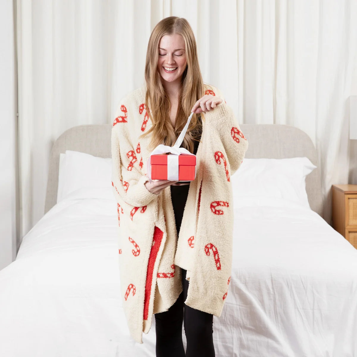 Woman wrapped in cream oversized blanket with red candy cane pattern, holding gift box on bed