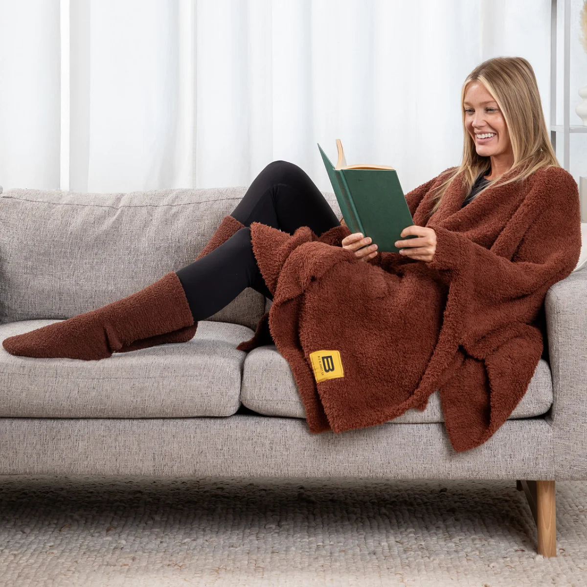 Woman relaxing on gray sofa wrapped in chocolate brown oversized plush blanket while reading, with matching socks