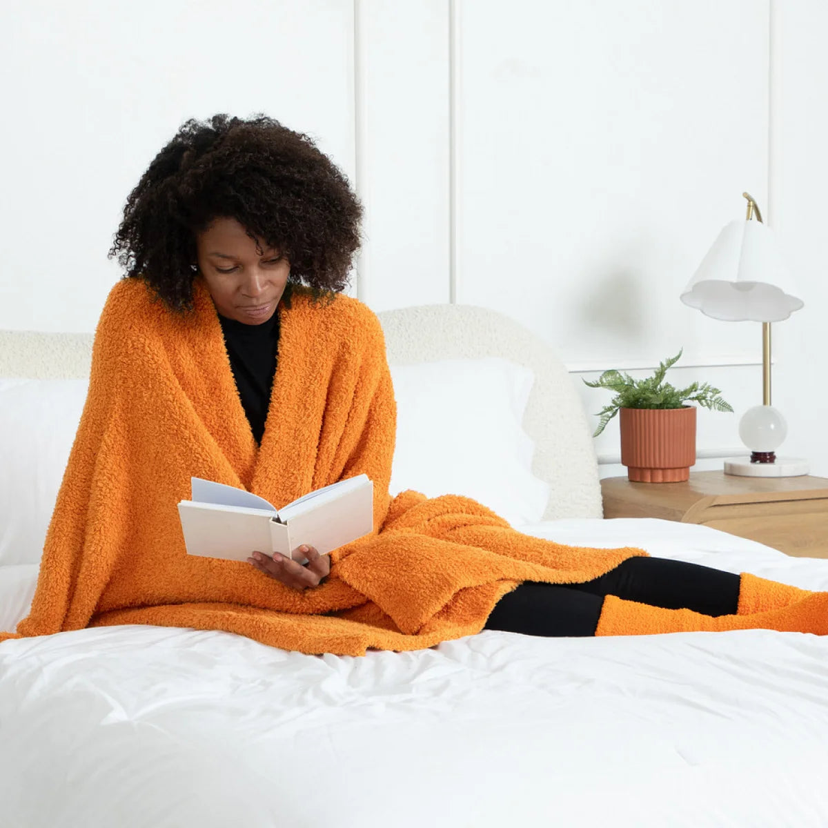 Woman wrapped in burnt orange oversized blanket reading on bed, Premier Plush Wrap shown in Fireside color