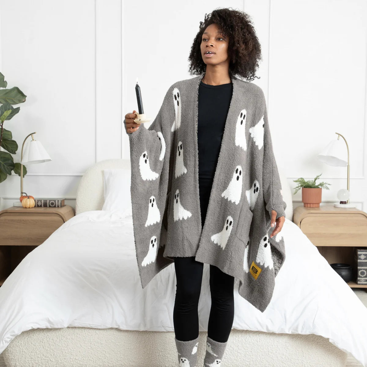 Woman wearing gray oversized blanket wrap with white ghost pattern, draped over shoulders in bedroom setting