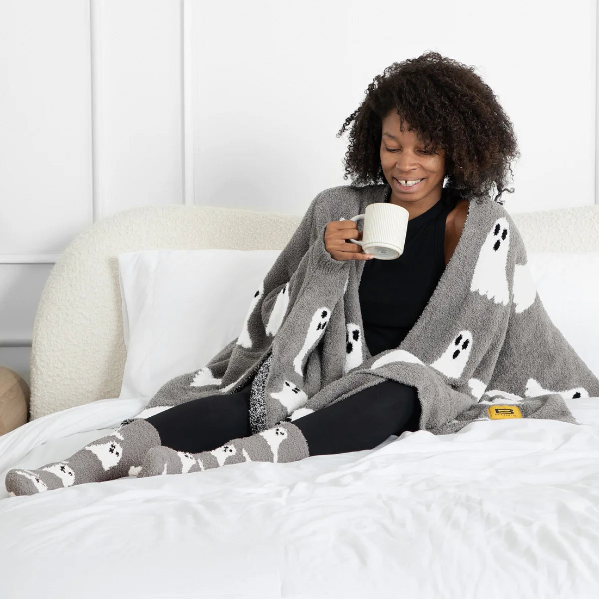 Woman on bed wearing gray oversized blanket with white ghost pattern, holding mug, paired with matching ghost socks